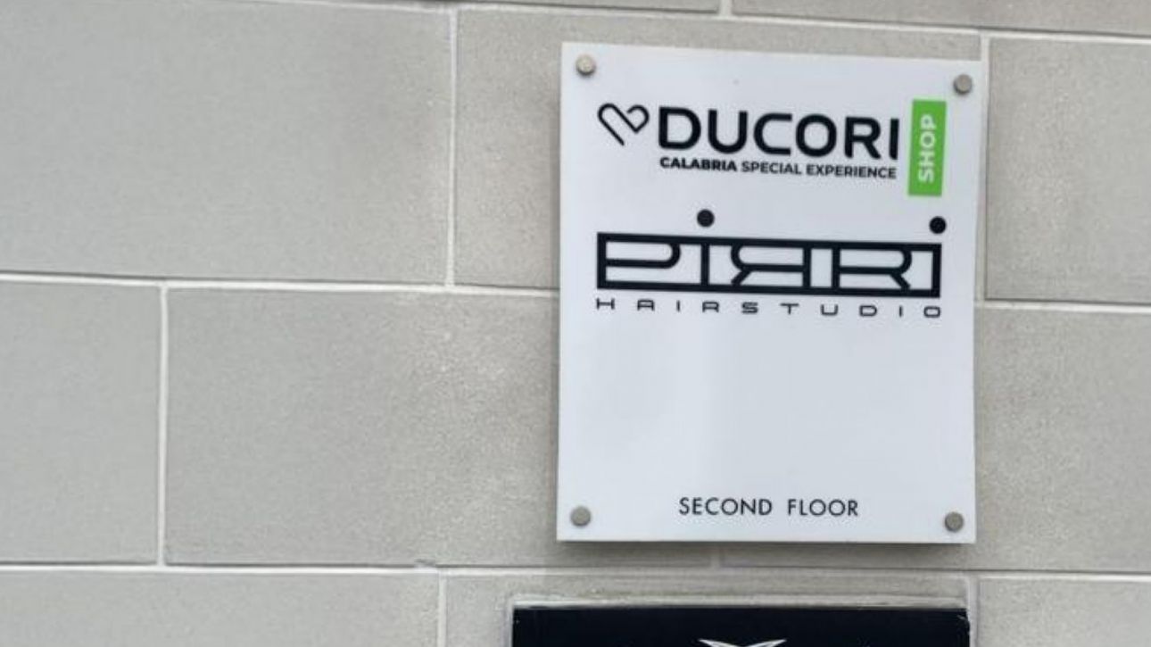 A wall-mounted sign displaying the "Ducori" logo with the tagline "Calabria Special Experience" and a green label reading "Shop." Below it, another sign reads "Pirri Hair Studio," indicating a second-floor location. The wall is made of light gray tiles.