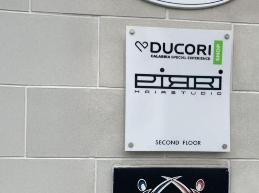 A wall-mounted sign displaying the "Ducori" logo with the tagline "Calabria Special Experience" and a green label reading "Shop." Below it, another sign reads "Pirri Hair Studio," indicating a second-floor location. The wall is made of light gray tiles.